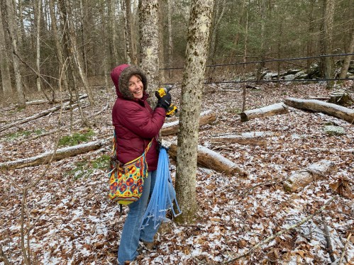 A person in a maroon jacket and colorful bag smiles while measuring a tree in a snowy forest.