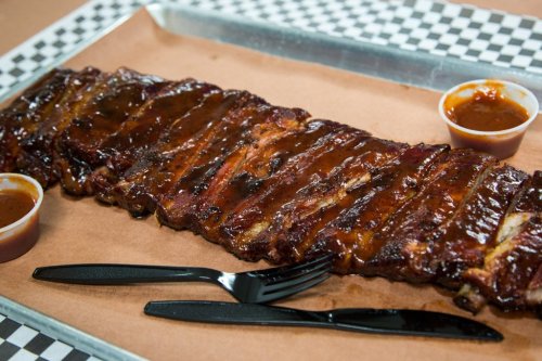 A platter of glazed barbecue ribs on brown paper, accompanied by two small cups of sauce and black utensils.