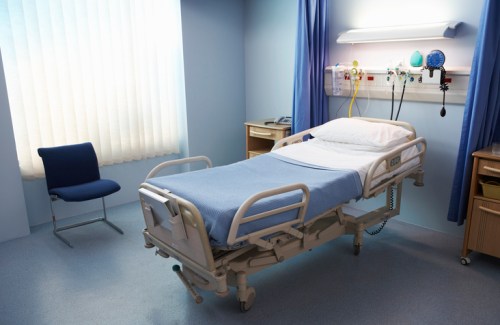 A hospital room featuring a bed with blue linens, a chair, and medical equipment on the wall.