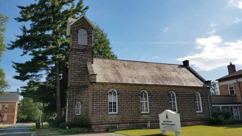 Historic stone church with a tall bell tower, surrounded by trees and a clear blue sky. Sign in front indicates its name.