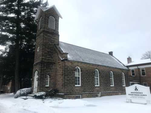 A stone church with a tall steeple, surrounded by snow, featuring a sign for Independence Presbyterian Church.