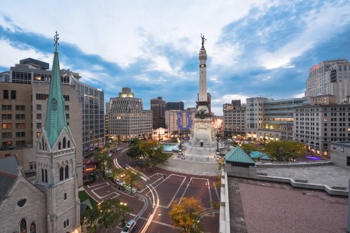 Aerial view of a city square featuring a monument, buildings, and a church steeple under a cloudy sky at dusk.