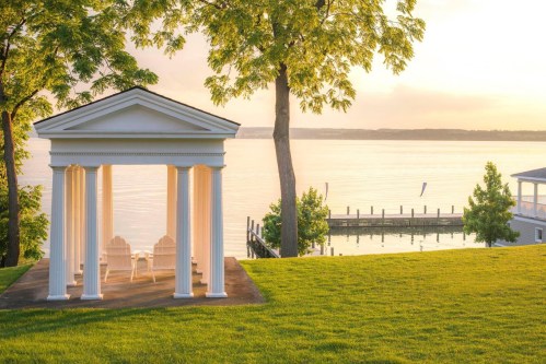 A serene lakeside scene featuring a white gazebo with columns, surrounded by lush greenery and calm water at sunset.