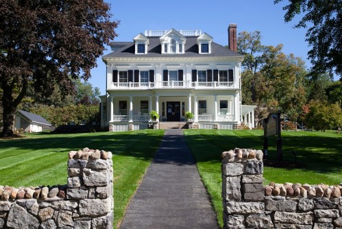 A large, elegant white house with a black roof, surrounded by green lawns and stone walls, under a clear blue sky.