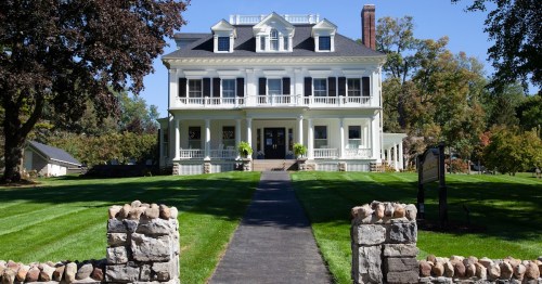 A large, elegant white mansion with a black roof, surrounded by green lawns and trees, with a stone entrance pathway.