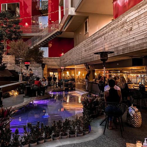 A vibrant indoor space with plants, a pool, and people socializing at a bar under warm lighting.