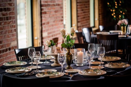 Elegant dining table set with plates, glasses, and floral centerpieces against a brick wall backdrop.