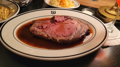 A large slice of prime rib served in a rich gravy, accompanied by side dishes in the background.