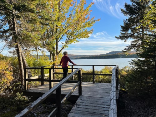 A person stands on a wooden deck overlooking a serene lake surrounded by trees and autumn foliage.