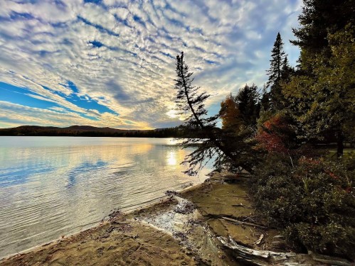 A serene lakeside scene at sunset, featuring clouds, trees, and calm water reflecting the colorful sky.