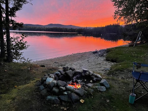 A serene lakeside scene at sunset, featuring a fire pit surrounded by rocks and a vibrant orange and purple sky.