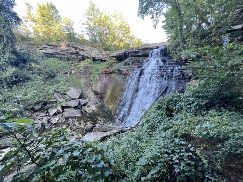 A serene waterfall cascades over rocky cliffs, surrounded by lush greenery and trees in a tranquil natural setting.