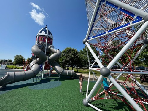 A playground featuring a rocket-shaped slide and a climbing structure, with children playing on green turf under a blue sky.