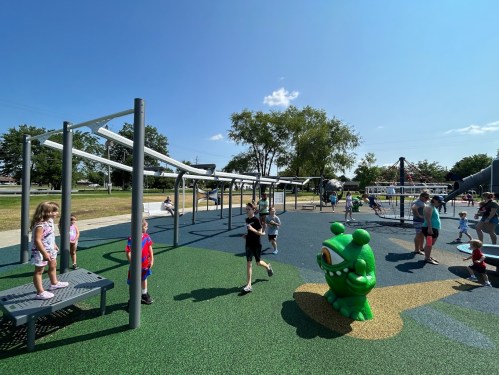Children play on a colorful playground with climbing structures and a large green frog sculpture under a clear blue sky.