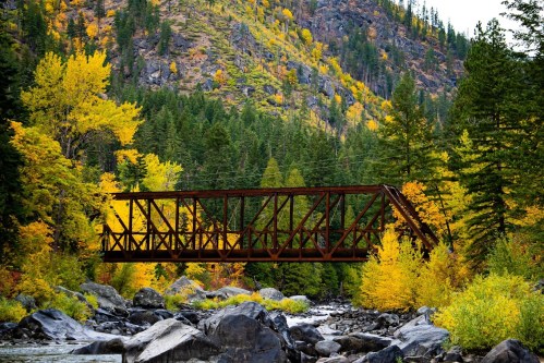 A rusted bridge spans a river, surrounded by vibrant autumn foliage and rocky terrain in a mountainous landscape.