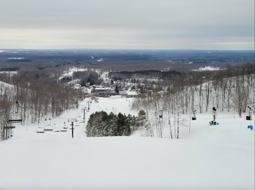 A snowy landscape with ski lifts and a view of a winter resort nestled among trees under a cloudy sky.