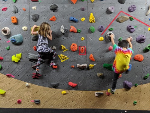 Two children climbing a colorful indoor rock wall, one in a striped shirt and the other in a tie-dye shirt.