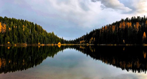 A serene lake surrounded by lush green and autumn-colored trees, reflecting the cloudy sky above.