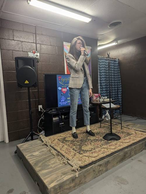 A person stands on a stage with a microphone, wearing a blazer and jeans, in a casual performance space.