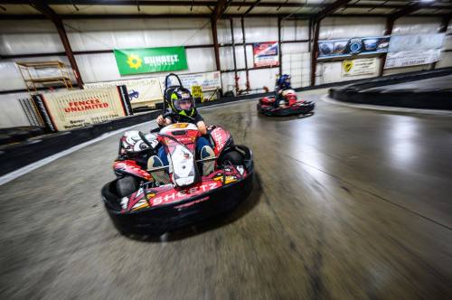Two go-karts racing on an indoor track, with one driver leaning into a turn. Bright banners decorate the walls.