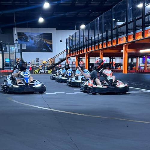 A group of go-karts racing on an indoor track, with spectators in the background.