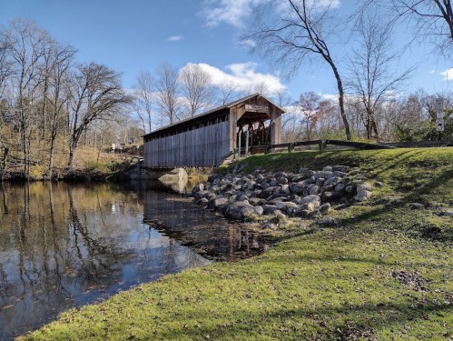A wooden covered bridge spans a calm river, surrounded by trees and rocky banks under a clear blue sky.