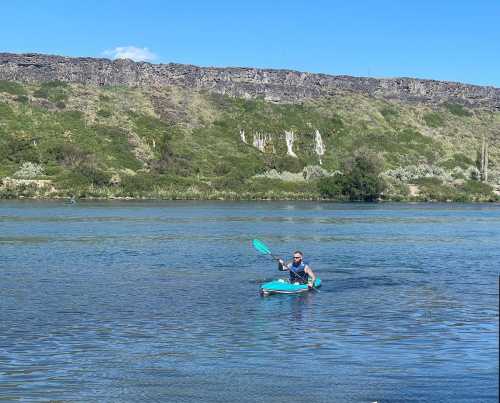 A person paddles a kayak on a calm river, surrounded by lush greenery and rocky cliffs under a clear blue sky.