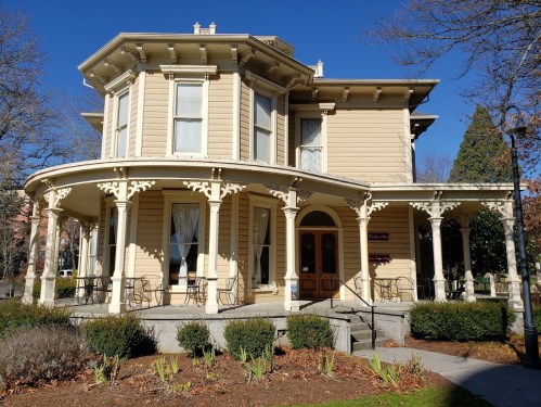 A historic Victorian-style house with a wraparound porch, surrounded by greenery and clear blue skies.