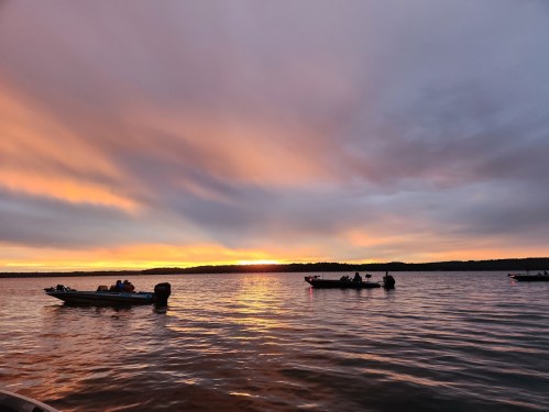 Boats on a calm lake at sunset, with vibrant orange and purple clouds reflecting on the water.
