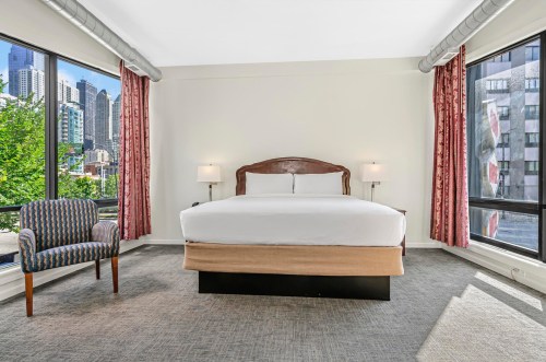 A bright hotel room featuring a king bed, armchair, and large windows with city views.