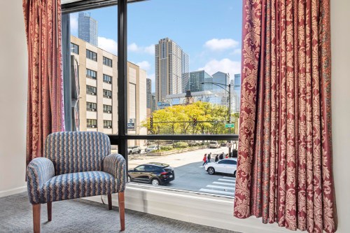 A cozy chair by a large window with a view of city buildings and a tree-lined street outside.