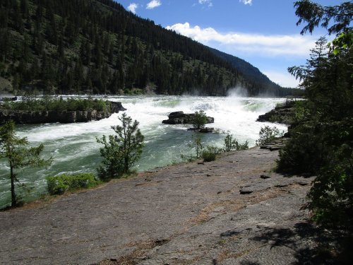 A rushing river flows through a rocky landscape, surrounded by lush green trees and mountains under a blue sky.