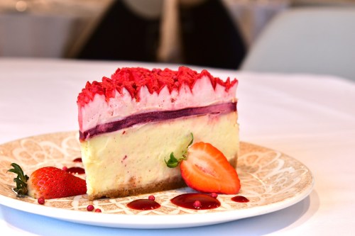 A slice of cheesecake with pink frosting, berry layers, and a fresh strawberry on a decorative plate.