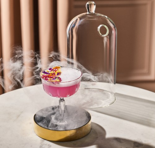 A pink cocktail with colorful garnishes, under a glass dome, emitting smoke, on a marble table.