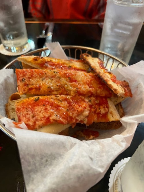 A basket of cheesy garlic bread topped with marinara sauce, served at a restaurant with drinks in the background.