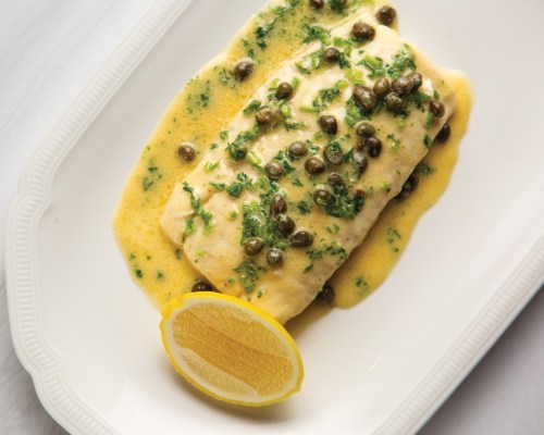 A plated fish dish topped with capers and herbs, drizzled with a creamy sauce, accompanied by a lemon wedge.