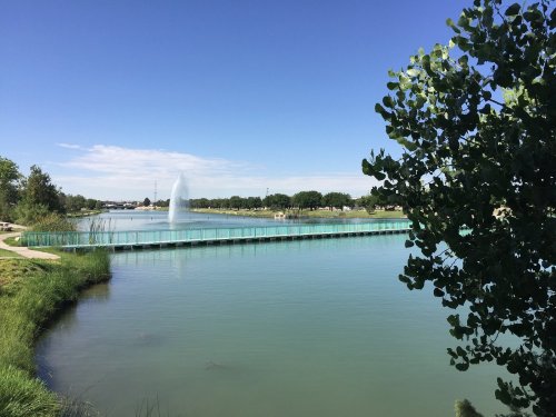 A serene lake with a fountain, surrounded by greenery and a wooden walkway under a clear blue sky.