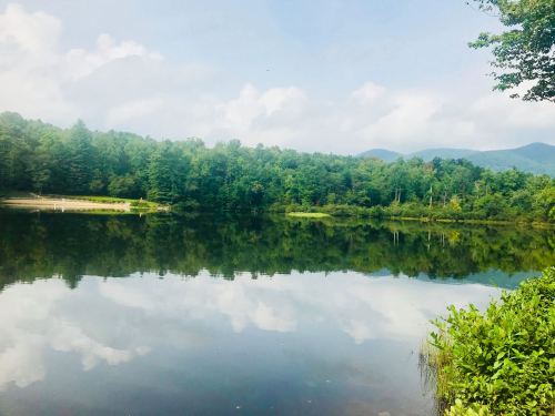 A serene lake surrounded by lush green trees, reflecting the sky and mountains in the calm water.