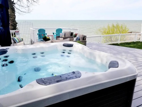 A hot tub on a deck overlooking a calm lake, with chairs and plants in the background.