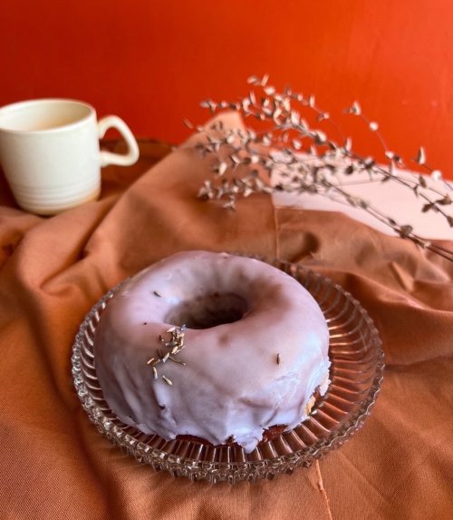 A lavender-glazed donut on a glass plate, with a cup and dried flowers in the background against an orange backdrop.