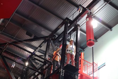 A person in safety gear stands on a platform, preparing to descend from a climbing structure with a harness and pulley system.