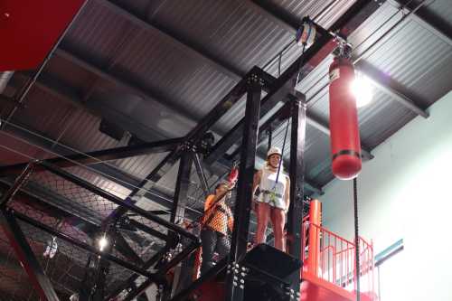 A person in safety gear stands on a platform, preparing to descend from a climbing structure with a harness and pulley system.