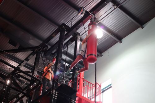 A person hangs from a red punching bag in a gym, while another person assists from below.