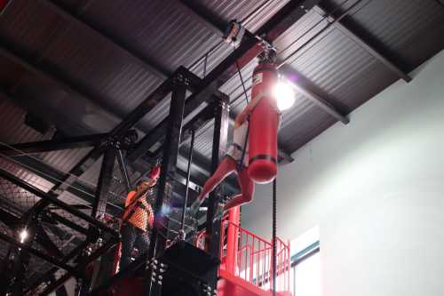 A person hangs from a red punching bag in a gym, while another person assists from below.