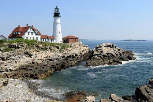 A lighthouse stands on rocky shores, with a coastal house nearby and a clear blue sky above.