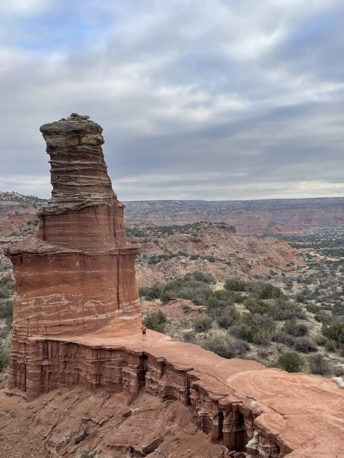 A lone hiker walks along a narrow path on a red rock formation, surrounded by rugged terrain and cloudy skies.