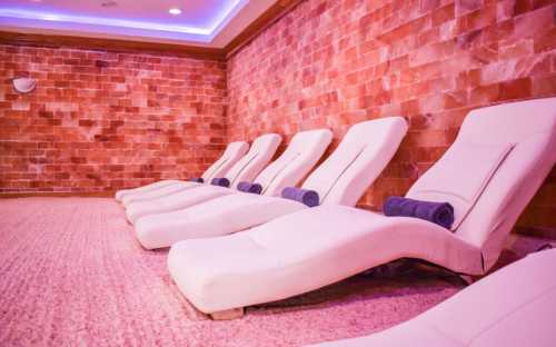 A serene spa room with white lounge chairs and a pink salt brick wall, creating a relaxing atmosphere.