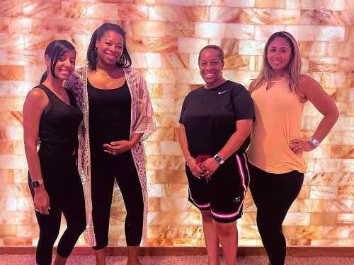 Four women stand together, smiling, in front of a softly lit salt wall. They are dressed in casual, comfortable clothing.