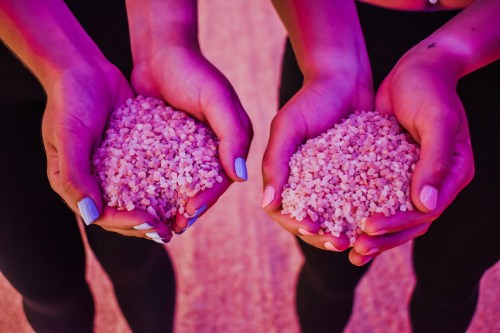 Two hands hold pink salt crystals, illuminated with purple lighting, against a soft, textured background.