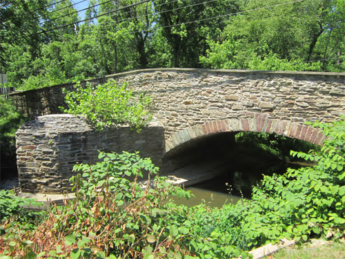 A stone bridge arches over a calm stream, surrounded by lush greenery and trees under a clear blue sky.